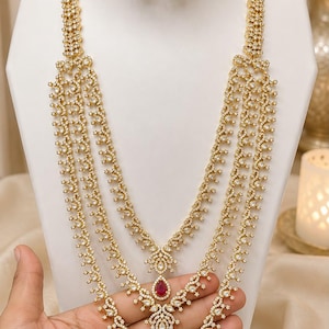 Pode incluir: Um colar dourado de várias camadas com padrões de filigrana intrincados e pedras transparentes, acentuado com três pedras vermelhas em forma de gota. O conjunto inclui brincos a condizer com um design semelhante de pedras e gotas vermelhas.