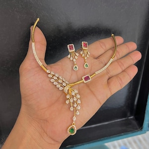 May include: A gold-toned necklace and earring set featuring clear pavé stones. The necklace has a curved bar design with a dangling leaf-like pendant accented by a green teardrop stone and a rectangular red stone. Matching earrings feature red stones.