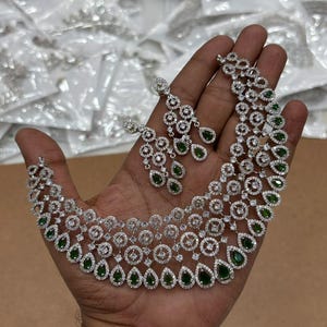 Puede incluir: Un conjunto de collar y pendientes en tono plateado que presenta múltiples filas de eslabones circulares incrustados con cristales transparentes y acentuados por piedras verdes colgantes en forma de pera. El conjunto muestra un diseño complejo.