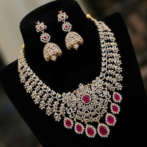 Op de afbeelding: Een sieradenset bestaande uit een ketting en bijpassende jhumka-oorbellen. De ketting heeft een centrale ovale robijn omringd door heldere stenen in een bloemmotief, met bungelende robijnen druppels en een rankachtig ontwerp. De oorbellen hebben een bijpassend bloemmotief met een centrale robijn en een klokvormige basis met een kleine parel.
