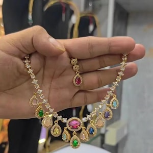 May include: A gold-tone necklace featuring a central oval pink gemstone surrounded by smaller teardrop-shaped stones in various colours including green, blue, yellow, and purple. The chain is accented with clear crystals and a matching earring.