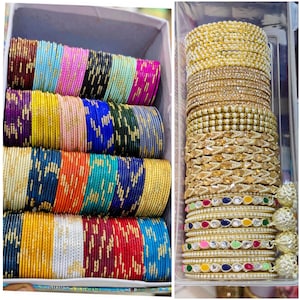 May include: A collection of multi-colored metal bangles arranged in rows within a box, featuring solid colors like red, blue, yellow, and pink with gold accents, alongside a separate container of gold-toned bangles with pearls and gems.