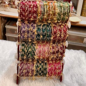 May include: A vertical display stand holding multiple sets of colorful bangles in shades of red, green, purple, and pink. Each bangle features a metallic gold finish with intricate patterns and small dangling gold charms attached to the sides.