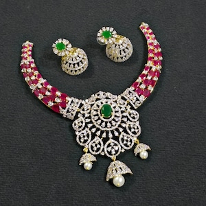 May include: A necklace and matching pair of jhumka earrings featuring a silver-toned metal base encrusted with clear crystals. The necklace has a central circular pendant with a green oval stone, red stones, and three dangling white pearls.