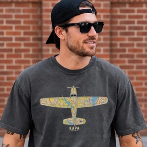 May include: A charcoal grey short-sleeve t-shirt featuring a graphic of a vintage aeroplane overlaid with a map pattern. The text KAPA is printed below the aeroplane illustration. The shirt has a crew neckline and a relaxed, washed-out fabric finish.