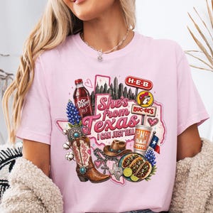 May include: A light pink t-shirt featuring a graphic print of Texas-themed icons including a Dr Pepper bottle, a cowboy boot, a hat, tacos, and logos for H-E-B and Buc-ee's, with the text "She's from Texas I CAN JUST TELL" in pink and white.