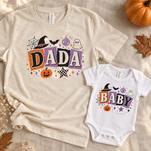 May include: A heather beige t-shirt and a white baby bodysuit featuring Halloween-themed graphics. The designs include the words DADA and BABY in block letters, surrounded by illustrations of a witch hat, ghost, pumpkin, bat, and disco ball.