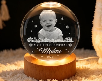 May include: A clear glass sphere night light featuring a laser-engraved portrait of a baby, snowflake patterns, and the text "MY FIRST CHRISTMAS Malou 2026". The sphere rests on a round, light-colored wooden base that illuminates the glass from below.