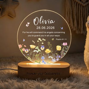May include: A circular acrylic night light with a wooden base featuring the name Olivia, the date 28.06.2026, the text For he will command his angels concerning you to guard you in all your ways Psalm 91:11, and colourful wildflower illustrations.