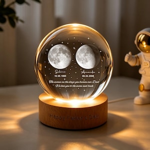 Può includere: Una lampada a sfera in vetro trasparente su una base rotonda in legno. La sfera presenta immagini incise al laser di due lune, i nomi Selina e Amanda, le date 19.05.1995 e 09.04.2008, e il testo The moons on the days you became our Dad. We love you to the moon and back.