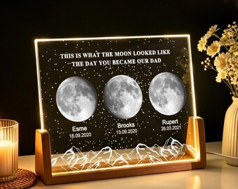 May include: A rectangular acrylic plaque with a light-up wooden base featuring three moon phases, a starry sky background, and mountain silhouettes. The text reads: THIS IS WHAT THE MOON LOOKED LIKE THE DAY YOU BECAME OUR DAD, Esme 18.09.2020, Brooks 13.09.2020, Rupert 26.03.2021.