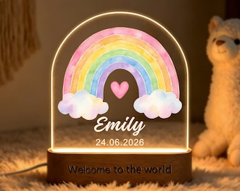 May include: An arched acrylic night light featuring a watercolor rainbow, a pink heart, and the text Emily 24.06.2026. The light sits on a wooden base engraved with the text Welcome to the world, emitting a warm glow from the bottom edge.