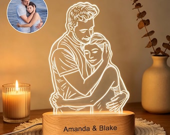 May include: A custom LED acrylic lamp featuring a white line-art illustration of a couple embracing, based on the circular photo inset. The clear acrylic panel sits in a light-colored wooden base engraved with the text Amanda & Blake.