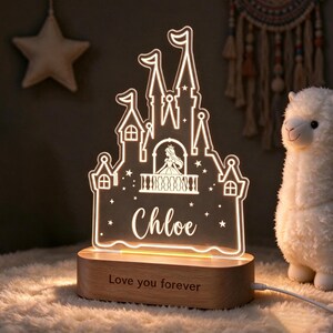 Puede incluir: Una luz nocturna de acr&iacute;lico transparente con forma de castillo, una silueta de princesa en un balc&oacute;n, el nombre Chloe y estrellas peque&ntilde;as. Se apoya en una base de madera clara con el texto grabado Love you forever.