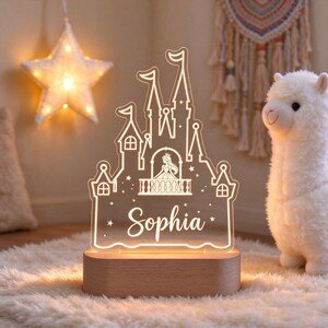 Puede incluir: Una luz nocturna de acr&iacute;lico transparente con forma de castillo y la silueta de una princesa en un balc&oacute;n. El nombre Sophia est&aacute; grabado en letra cursiva debajo del castillo. La pieza est&aacute; montada sobre una base de madera clara ovalada.