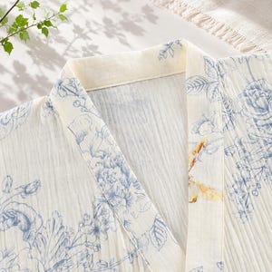 May include: A cream-colored robe featuring a blue floral print and a single yellow dragonfly detail on the collar. The garment has a V-neckline, a structured lapel, and a textured, crinkled fabric finish throughout the entire piece.