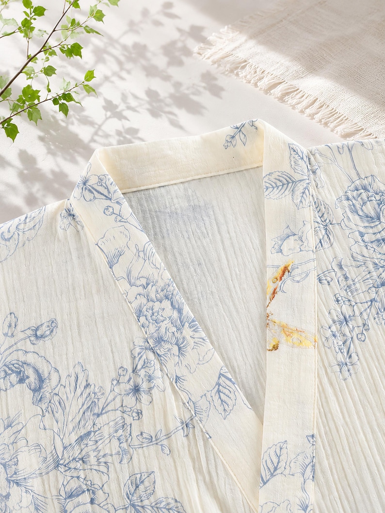 May include: A cream-colored robe featuring a blue floral print and a single yellow dragonfly detail on the collar. The garment has a V-neckline, a structured lapel, and a textured, crinkled fabric finish throughout the entire piece.