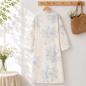May include: A long-sleeved, knee-length robe in an off-white fabric featuring a blue and yellow floral print. The garment has a simple collar, a straight hemline, and is displayed on a wooden hanger against a plain wall.