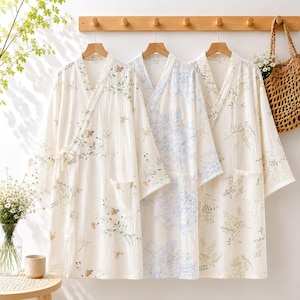 May include: Three long-sleeved robes made of white textured fabric, each featuring a different botanical print. The left robe has a butterfly and leaf pattern, the centre has a blue floral design, and the right robe displays a green fern motif.