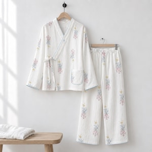Op de afbeelding: Een witte pyjamaset met lange mouwen, bestaande uit een wikkeltop met een striksluiting aan de zijkant, een opgestikte zak en een bijpassende broek met wijde pijpen. De stof heeft een textuur en een herhalend bloemenpatroon in roze en blauw.