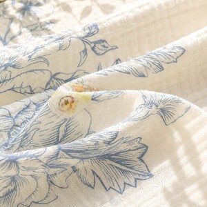 May include: A white cotton muslin fabric featuring a blue botanical print of leaves and flowers. The textile has a crinkled, multi-layered texture with soft folds and a subtle pattern of blue line-drawn foliage across the surface.