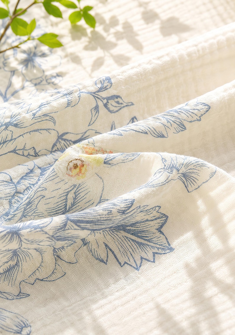 May include: A white cotton muslin fabric featuring a blue botanical print of leaves and flowers. The textile has a crinkled, multi-layered texture with soft folds and a subtle pattern of blue line-drawn foliage across the surface.
