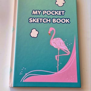 Puede incluir: Un cuaderno de bocetos de tapa dura color verde azulado con el texto MY POCKET SKETCH BOOK impreso en azul. La portada presenta un flamenco rosa de pie sobre una pata, dos nubes blancas y un diseño de onda rosa en la parte inferior con pequeños puntos blancos.