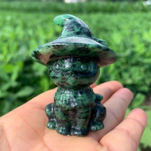 May include: A small, carved stone figurine of a cat wearing a pointed witch hat. The sculpture is crafted from a dark green mineral with black mottled patterns throughout, featuring smooth, polished surfaces and defined facial features.