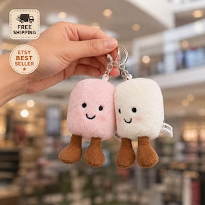 Könnte beinhalten: Ein Paar Plüsch-Schlüsselanhänger in Marshmallow-Form, einer in Hellrosa und einer in Weiß, jeweils mit einem lächelnden Gesicht, gestickten schwarzen Augen und Mund, rosa Wangen und zwei kleinen braunen Beinen aus Kord an einem Metallring.