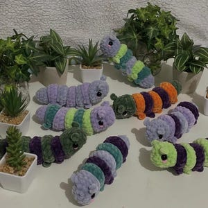 May include: A collection of plush caterpillar toys made from soft, textured yarn in various colour combinations including purple, green, orange, and blue. Each caterpillar features small dark eyes and a segmented body design, arranged among several small potted plants.