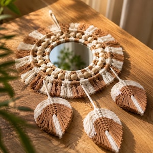 Könnte beinhalten: Ein runder Wandspiegel, der mit aufwendigem Makramee-Garn in abwechselnden Beige- und Cremetönen umrandet ist. Das Design umfasst einen Ring aus glatten Holzperlen, einen Fransenrand und drei hängende, blattförmige Makramee-Elemente.