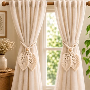 May include: Two off-white textured curtains hanging from a wooden rod, each held back by a macrame tieback featuring a central wooden bead and two leaf-shaped fringe accents. The curtains have a pleated top and a semi-sheer, woven fabric appearance.