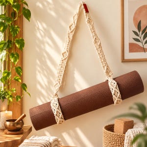 May include: A dark brown yoga mat suspended by a cream-colored macrame strap. The strap features intricate knotted diamond patterns and a small red rectangular tag at the top. The mat has a textured surface and is held securely by two loops.