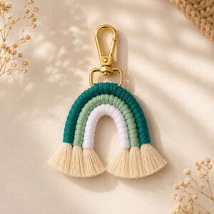Puede incluir: Un llavero de arcoíris de macramé con tres capas arqueadas en cordón de algodón verde oscuro, verde salvia y blanco. Los extremos del arcoíris terminan en flecos deshilachados de color crema. Un mosquetón de metal dorado está unido a la parte superior.