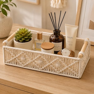 May include: A rectangular storage tray featuring a cream-coloured macrame exterior with a diamond pattern and light wood handles. Inside the tray are a small potted succulent, a glass perfume bottle, a candle, a reed diffuser, and a wristwatch.