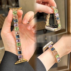 May include: A gold-toned watch strap featuring a series of rectangular, multi-coloured faceted crystals in shades of green, purple, blue, and peach. The strap is designed with metallic links and connects to a watch face, shown worn on a wrist.