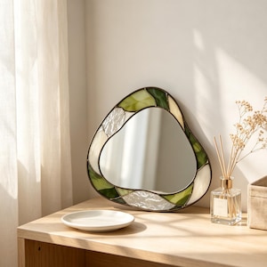 May include: An irregular, organic-shaped mirror framed in stained glass. The border features a combination of textured clear glass, opaque cream glass, and various shades of green glass pieces joined by dark metal solder lines.