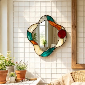 Könnte beinhalten: Ein Wandspiegel mit einem unregelmäßigen, wellenförmigen Rahmen aus Buntglasstücken in den Farben Petrol, Bernstein, Creme und einem kreisförmigen roten Akzent. Die Glassegmente sind durch dunkle Metalllinien verbunden, was eine fließende Form ergibt.