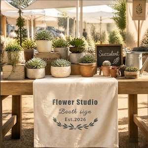 May include: A wooden table displays various potted succulents in ceramic and terracotta pots. A beige fabric runner features the text "Flower Studio Booth sign Est. 2026" with a leaf graphic. A small chalkboard sign reads "Succulent" next to a metal watering can.