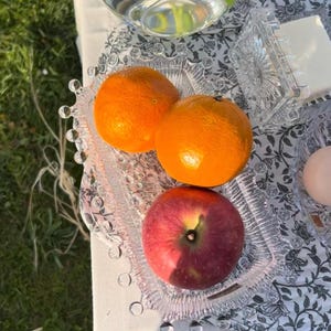 May include: A rectangular clear glass tray with a beaded edge holds two bright orange citrus fruits and one red apple. The tray sits on a white tablecloth featuring a black floral pattern, positioned near a glass vase and a glass container.