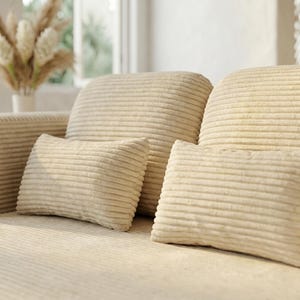 May include: A beige corduroy sofa featuring a textured, ribbed fabric surface. The sofa includes two rectangular throw pillows made of the same matching beige corduroy material, positioned against the back cushions of the seating furniture.