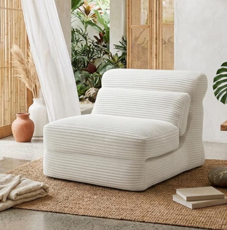 May include: A low-profile armless chair upholstered in white corduroy fabric with a distinct horizontal ribbed texture. The chair features a rounded backrest and a thick, cushioned seat, creating a singular, cohesive piece of furniture.