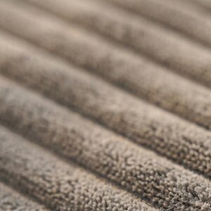 May include: A close-up view of a taupe-coloured fabric featuring a distinct ribbed texture. The material displays parallel, raised lines that run diagonally across the frame, showcasing a soft, fuzzy surface with a consistent, neutral tone throughout.