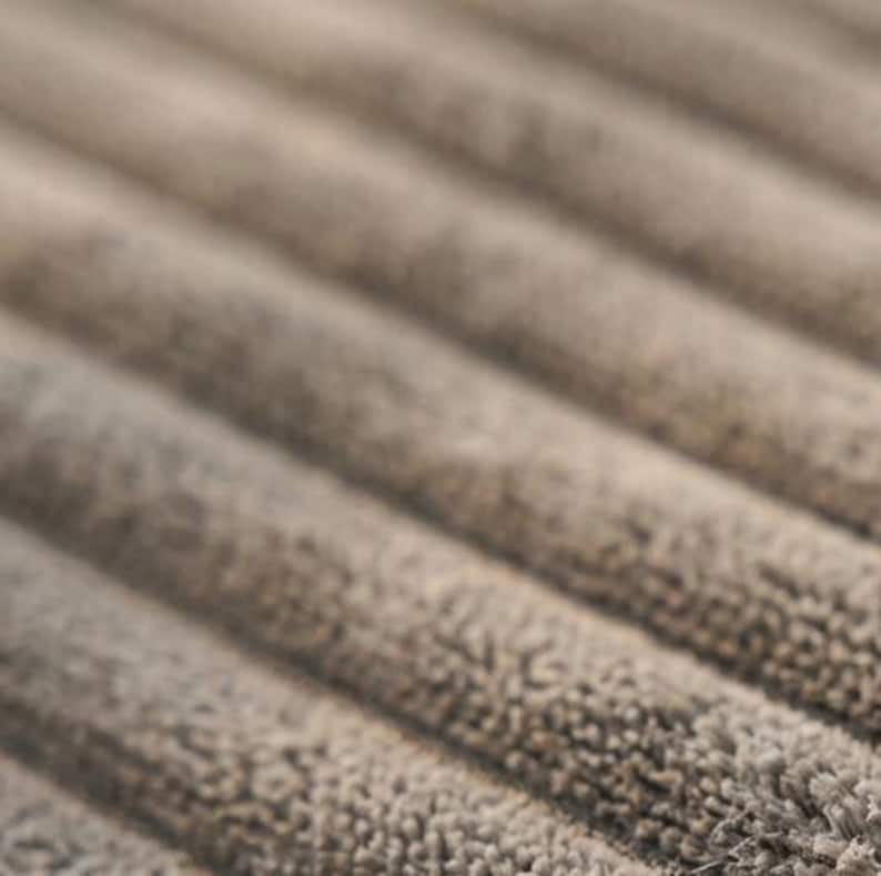 May include: A close-up view of a taupe-coloured fabric featuring a distinct ribbed texture. The material displays parallel, raised lines that run diagonally across the frame, showcasing a soft, fuzzy surface with a consistent, neutral tone throughout.