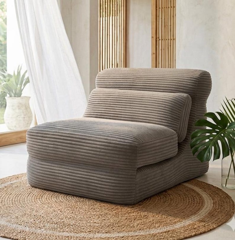 May include: A low-profile armless chair upholstered in a taupe-coloured corduroy fabric with a distinct horizontal ribbed texture. The chair features a rounded backrest, a separate horizontal lumbar cushion, and a thick, deep seat cushion.