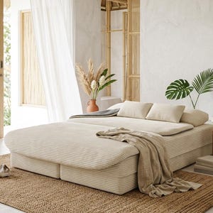 May include: A low-profile platform bed upholstered in cream-colored corduroy fabric. The bed features a matching corduroy duvet, two rectangular pillows, and a draped beige linen throw blanket, all set upon a textured woven jute area rug.