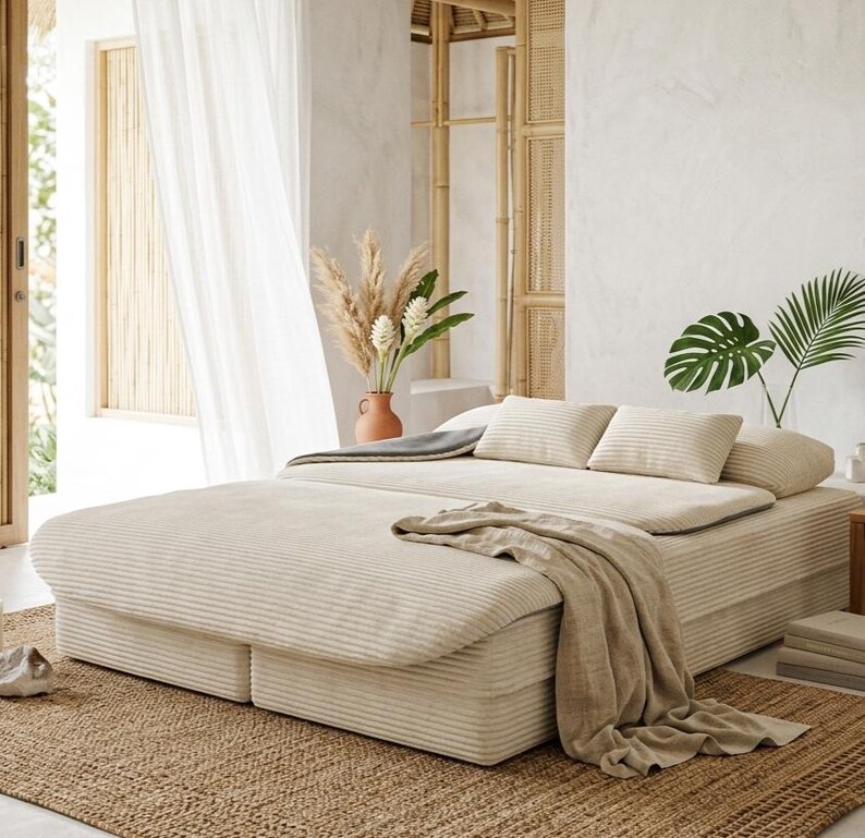 May include: A low-profile platform bed upholstered in cream-colored corduroy fabric. The bed features a matching corduroy duvet, two rectangular pillows, and a draped beige linen throw blanket, all set upon a textured woven jute area rug.