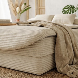 May include: A beige corduroy bed frame and mattress set featuring a horizontal ribbed texture. The bed is covered with a matching beige corduroy duvet and a draped, textured beige throw blanket. Two beige corduroy pillows rest at the head.