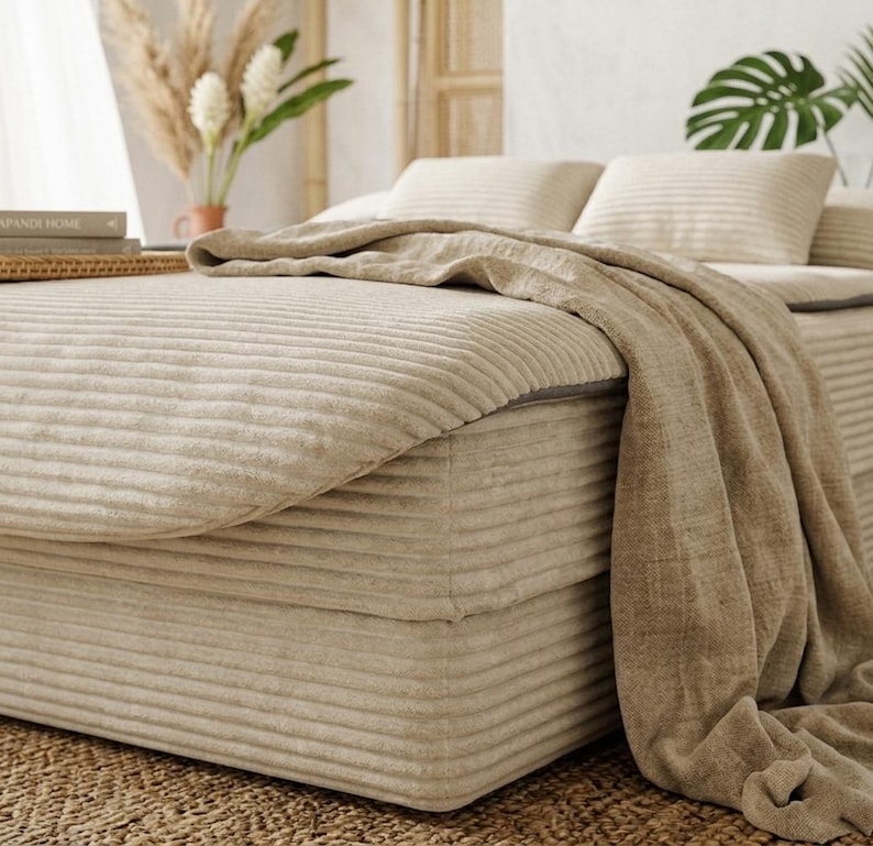 May include: A beige corduroy bed frame and mattress set featuring a horizontal ribbed texture. The bed is covered with a matching beige corduroy duvet and a draped, textured beige throw blanket. Two beige corduroy pillows rest at the head.