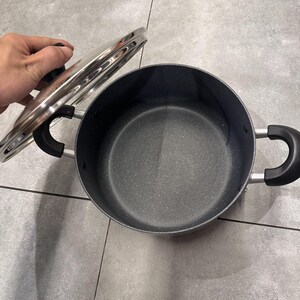 May include: A round cooking pot with a dark gray speckled non-stick interior and two black side handles. The pot includes a clear glass lid with a silver metal rim and a black knob handle, shown partially lifted to reveal the interior basin.